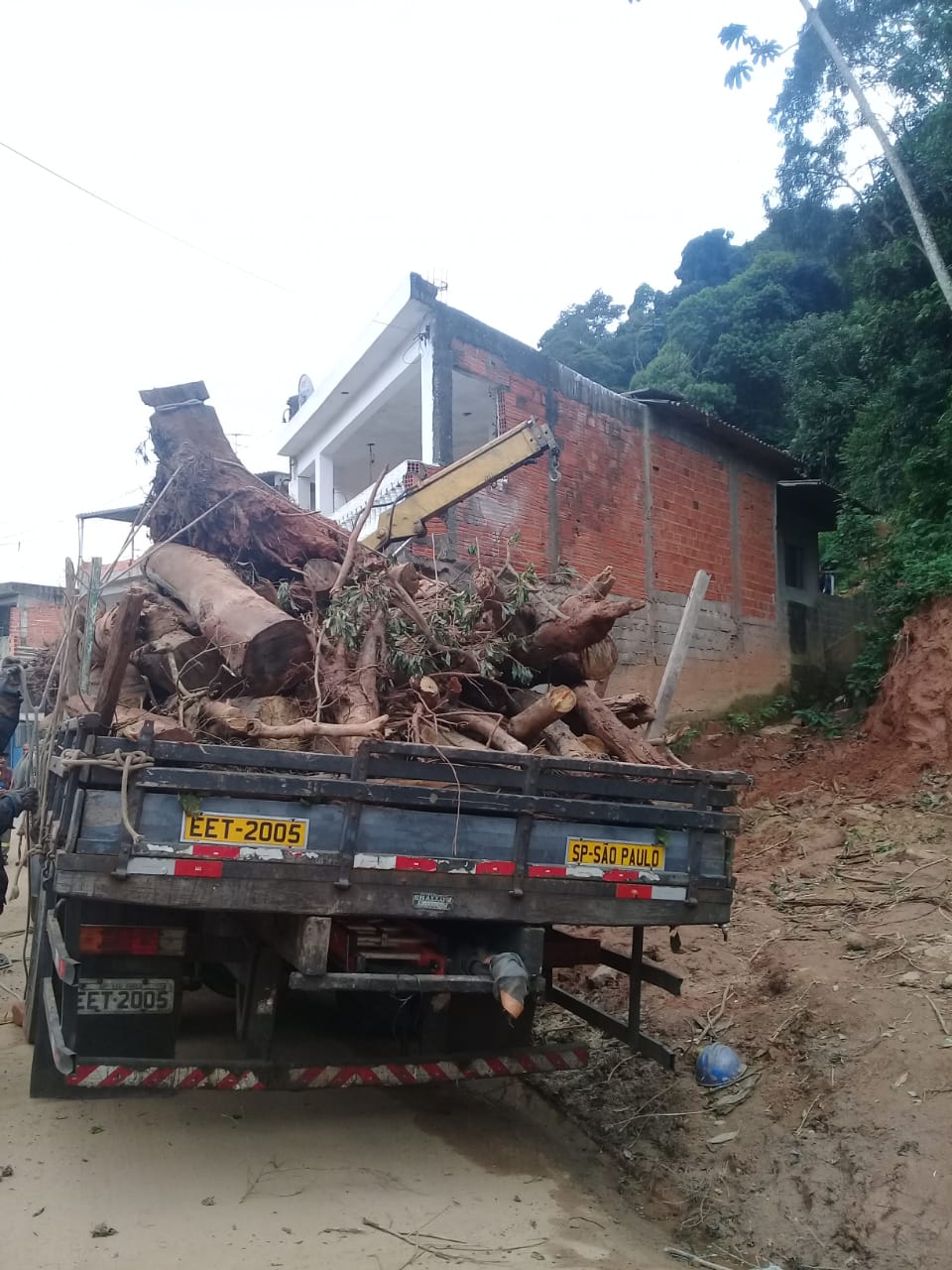 Caminhão está com a carroceria lotada de troncos e galhos de árvores, retirados de terreno onde houve deslizamento. Ao fundo, uma casa com a parede externa sem reboco, na rua Miguel Fernandes.